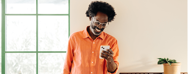 A man stands against a beige wall and smiles down at his smartphone. To his right is a window, and a plant is to his left.