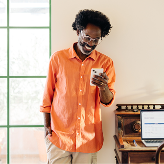 A man stands against a beige wall and smiles down at his smartphone. To his right is a window, and a plant is to his left.