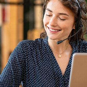 A woman wears a headset and takes down notes while sitting at her laptop. She smiles as she writes.