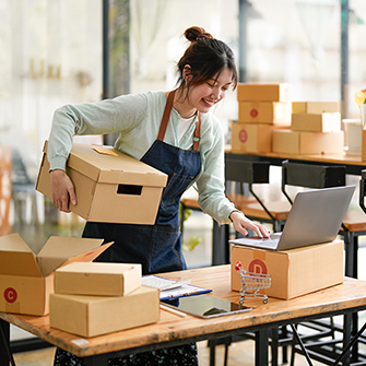 A woman surrounded by stacks of boxes looks into her laptop screen.