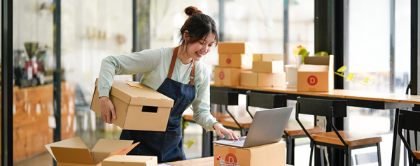 A woman surrounded by stacks of boxes looks into her laptop screen.