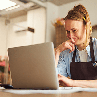 A person in an apron sits in front of a laptop looking at the screen.