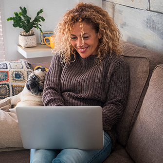A woman sits on a couch with a pug and her laptop.