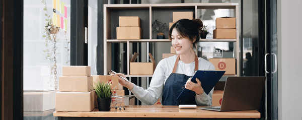 A woman holding a clipboard surrounded by a laptop and a stack of boxes.
