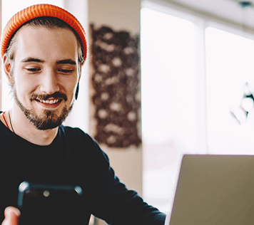 A man in a beanie sits in front of a laptop looking down at his smartphone.