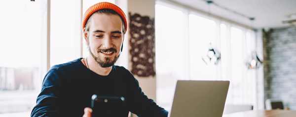 A man in a beanie sits in front of a laptop looking down at his smartphone.