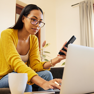 A woman in a yellow sweater and glasses sits in front of a laptop holding a smartphone.
