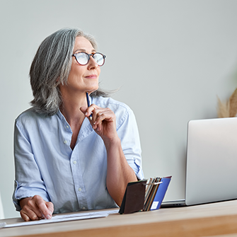 A woman with glasses looks into the distance while seated in front of a laptop.