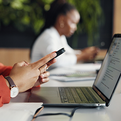 This image shows someone holding their smartphone sitting in front of a laptop and a blurry figure staring at their smartphone.