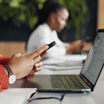 This image shows someone holding their smartphone sitting in front of a laptop and a blurry figure staring at their smartphone.