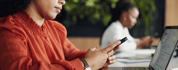 This image shows two women looking at their phone. The one upfront has a laptop in front of her.