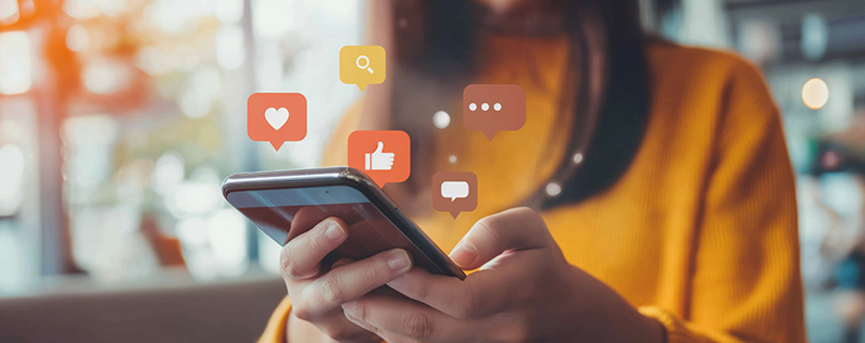 Header_Audengage A woman wearing a yellow sweater holds a smartphone. Social media engagement icons rise from the screen and float above it.