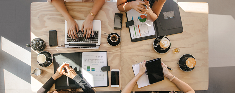 Header_Conversions Overhead view of a group of businesswomen sitting at a table to work together. Only the table and their arms are visible.