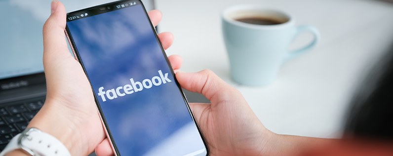 Header_FacebookMarketers A person sits at a desk and views the Facebook logo screen on their smartphone. A laptop and a cup of coffee are on the desk.