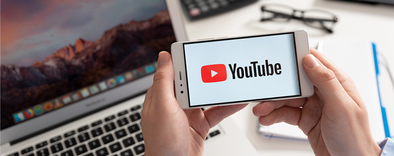 A smartphone displaying the YouTube logo. A person holds the phone above a desk with a laptop and a coffee mug.