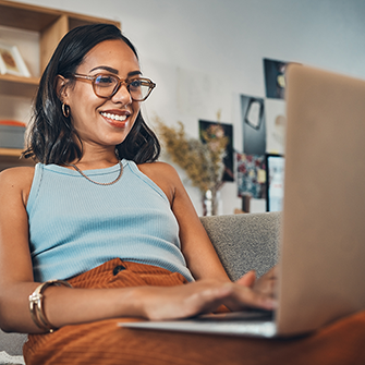 A smiling woman sits on a couch with her laptop on her lap.