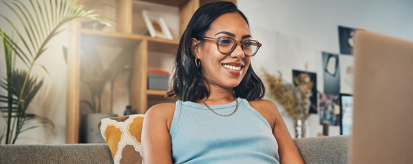A smiling woman sits on a couch with her laptop on her lap.
