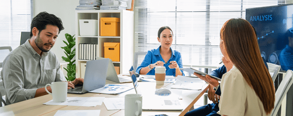 Four people sit at a table to talk. Documents, laptops, and coffee cups are on the table, and a television screen displays data charts.
