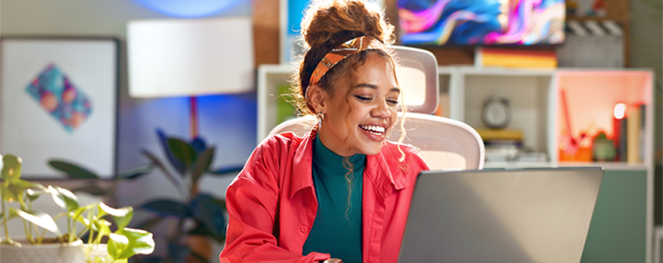 A woman sits at her laptop and smiles as she works. She wears a hot pink blazer and her office is decorated with colorful art.
