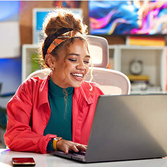 A woman sits at her laptop and smiles as she works. She wears a hot pink blazer and her office is decorated with colorful art.