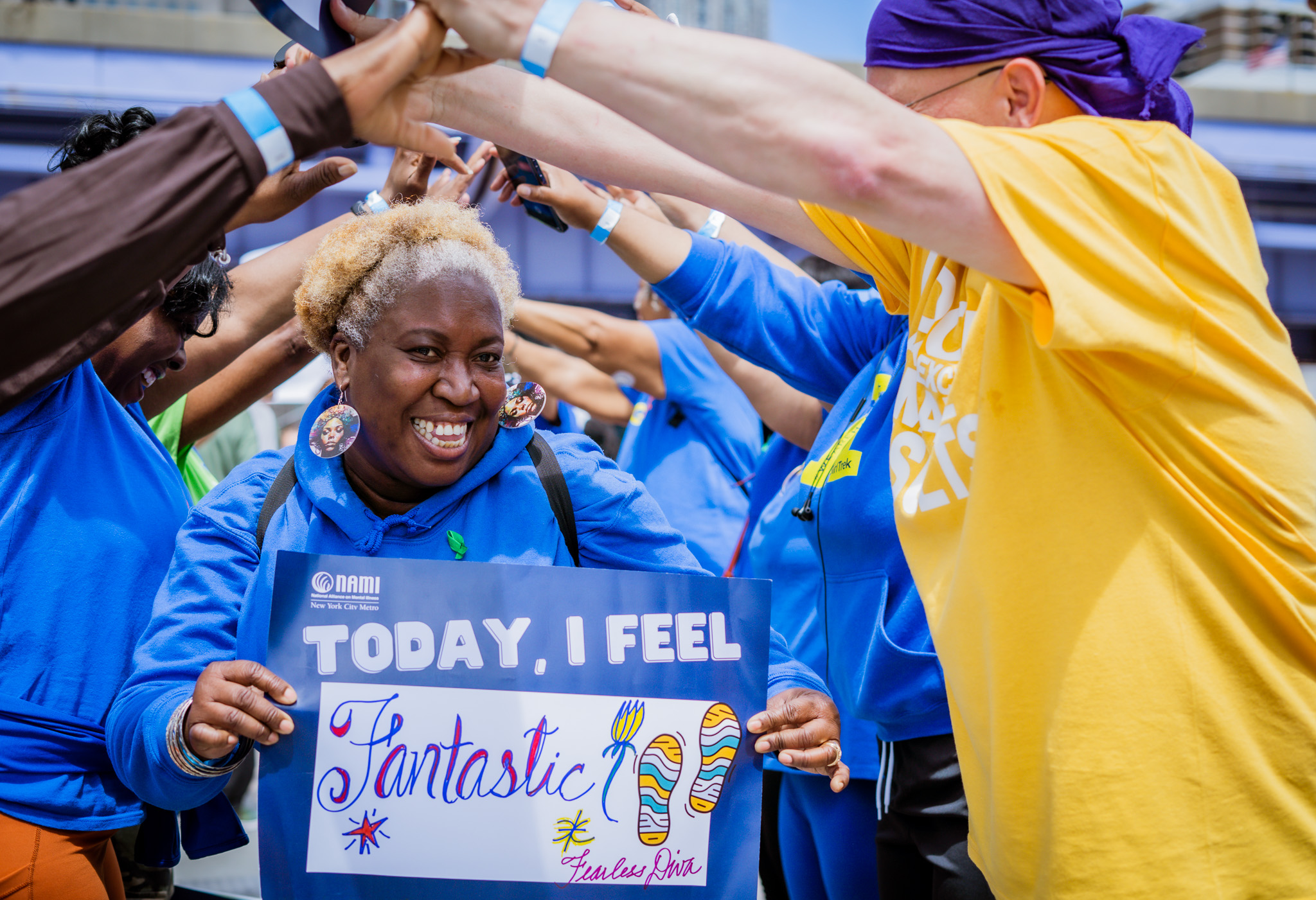 Smiling woman holding a sign that says “Today, I Feel Fantastic” while walking through a human arch of raised arms at a NAMI-NYC event.
