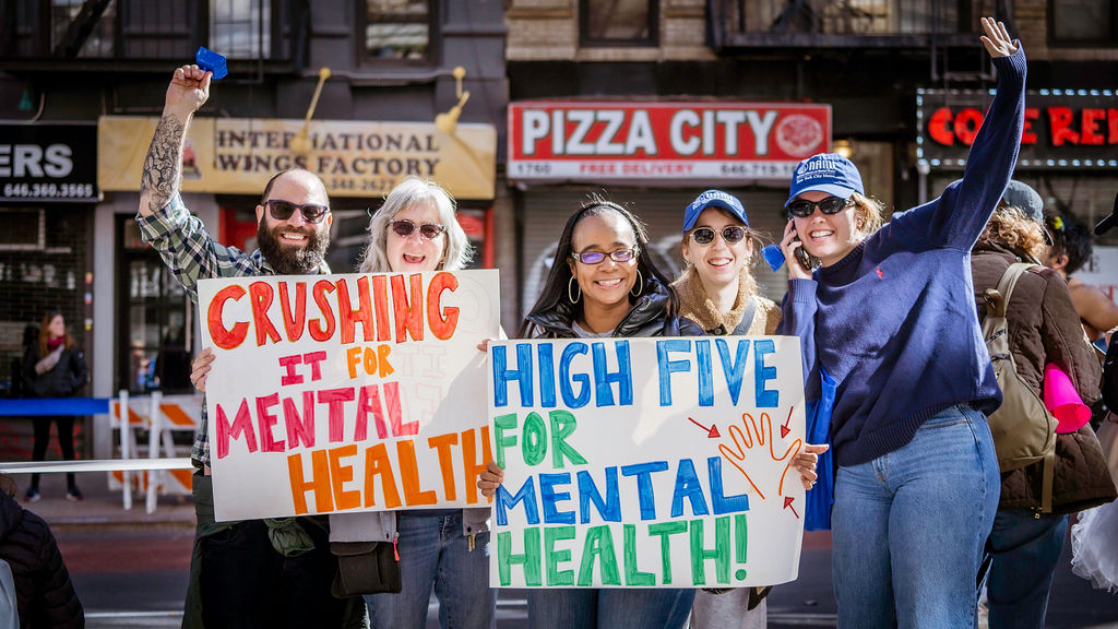 Group of five smiling people holding colorful signs that say “Crushing it for Mental Health” and “High Five for Mental Health” on a city street.