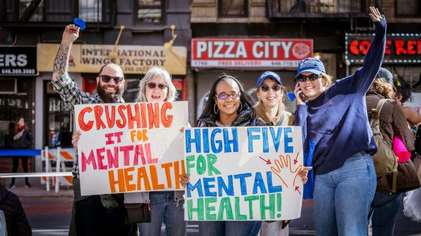Group of five smiling people holding colorful signs that say “Crushing it for Mental Health” and “High Five for Mental Health” on a city street.