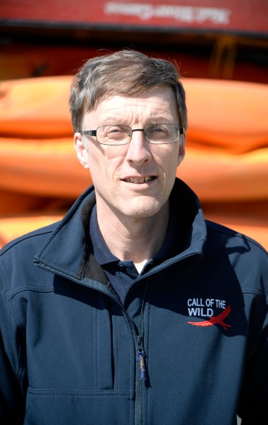 Mark Soanes, Director of Call of the Wild, wearing a navy jacket with the company logo, standing outdoors in front of orange kayaks.