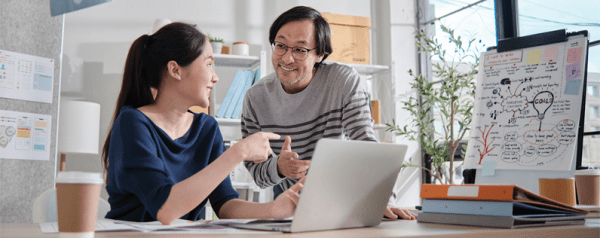 A man and a woman review documents on a laptop. They're in a brightly lit office, and a large notepad on an easel is behind them.