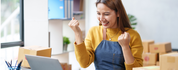A woman sits at her laptop and cheers after winning back a subscriber. A pile of shipping boxes is behind her.