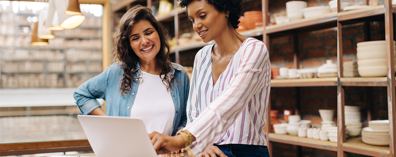 Header_SEOEmailMarketing Two women stand at a desk and review documents on a laptop. They stand in front of shelving units filled with pottery.