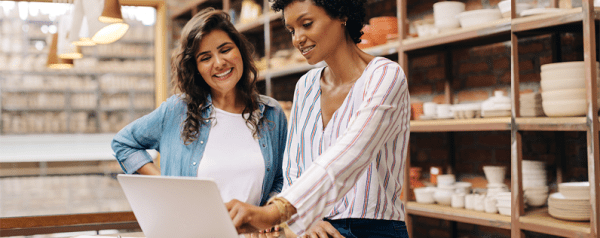 Two women stand at a desk and review documents on a laptop. They stand in front of shelving units filled with pottery.