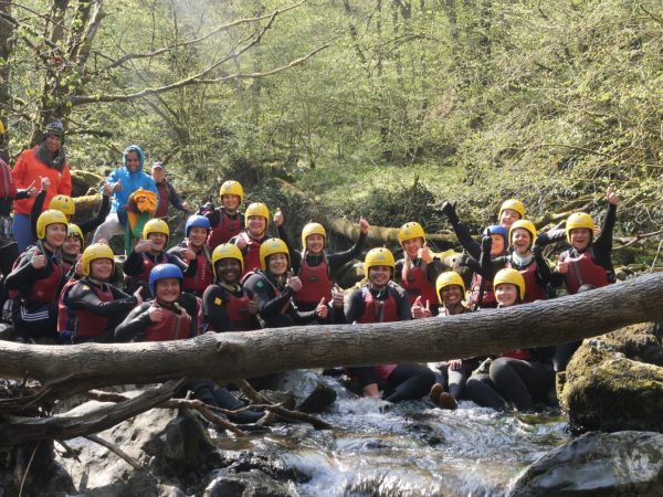 A large group of people in kayaking gear sitting in a river, smiling and posing for a group photo during an outdoor team-building activity.