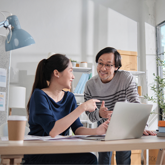 A man and a woman review documents on a laptop. They're in a brightly lit office, and a large notepad on an easel is behind them.