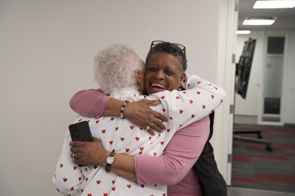 Two women warmly hugging in a bright room. One wears a white shirt with red hearts and holds a phone, while the other, smiling broadly, wears a pink top and black vest.