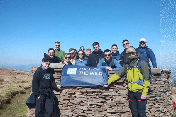 A small group of people in hiking attire smiling holding a navy blue banner with white text reading Call of the Wild with a company logo
