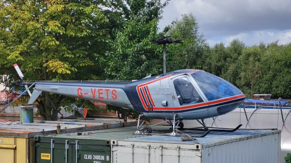 A light gray helicopter with red and black detailing, labeled &ldquo;G-VETS,&rdquo; displayed outdoors on top of shipping containers.