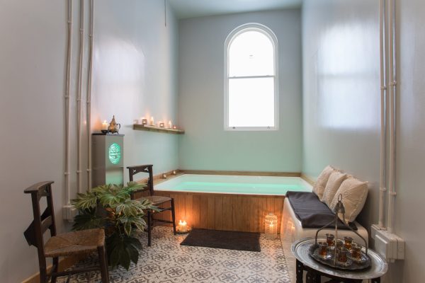 A softly lit spa room with a glowing float pool, candles, wooden chairs, patterned floor tiles, and a cushioned bench with pillows.