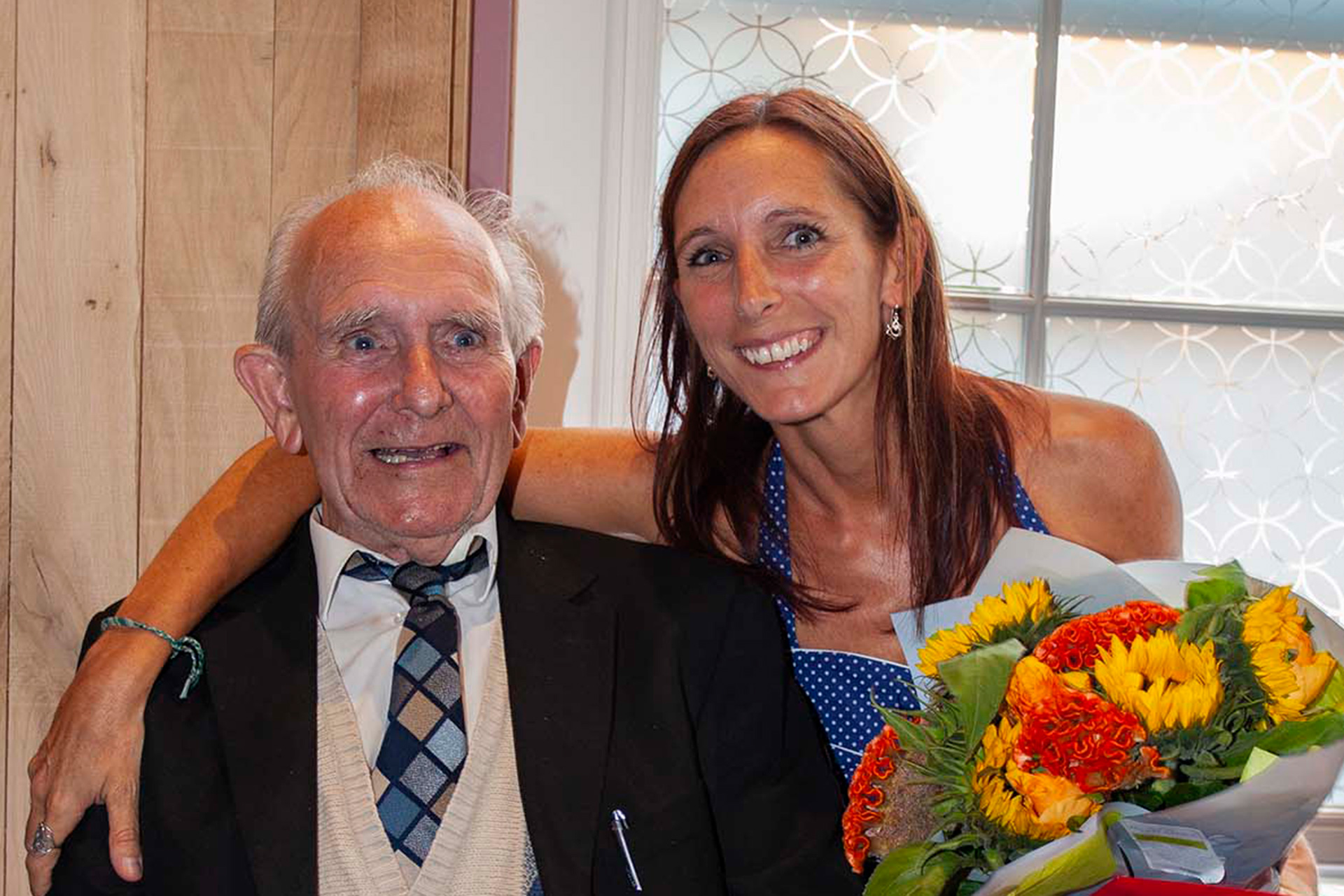 A smiling woman holding a bouquet of sunflowers embraces an elderly man in a suit and tie, both looking joyful together.