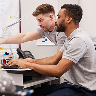 Two men sit together at a desk as one points to a computer screen.