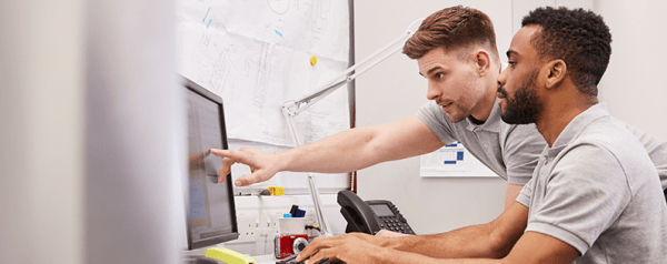 Two men sit together at a desk as one points to a computer screen.