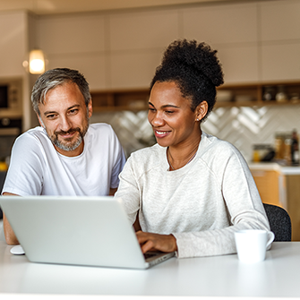 A man and a woman seated at a kitchen table look at a laptop screen.