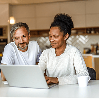 A man and a woman seated at a kitchen table look at a laptop screen.