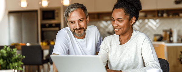 A man and a woman seated at a kitchen table look at a laptop screen.