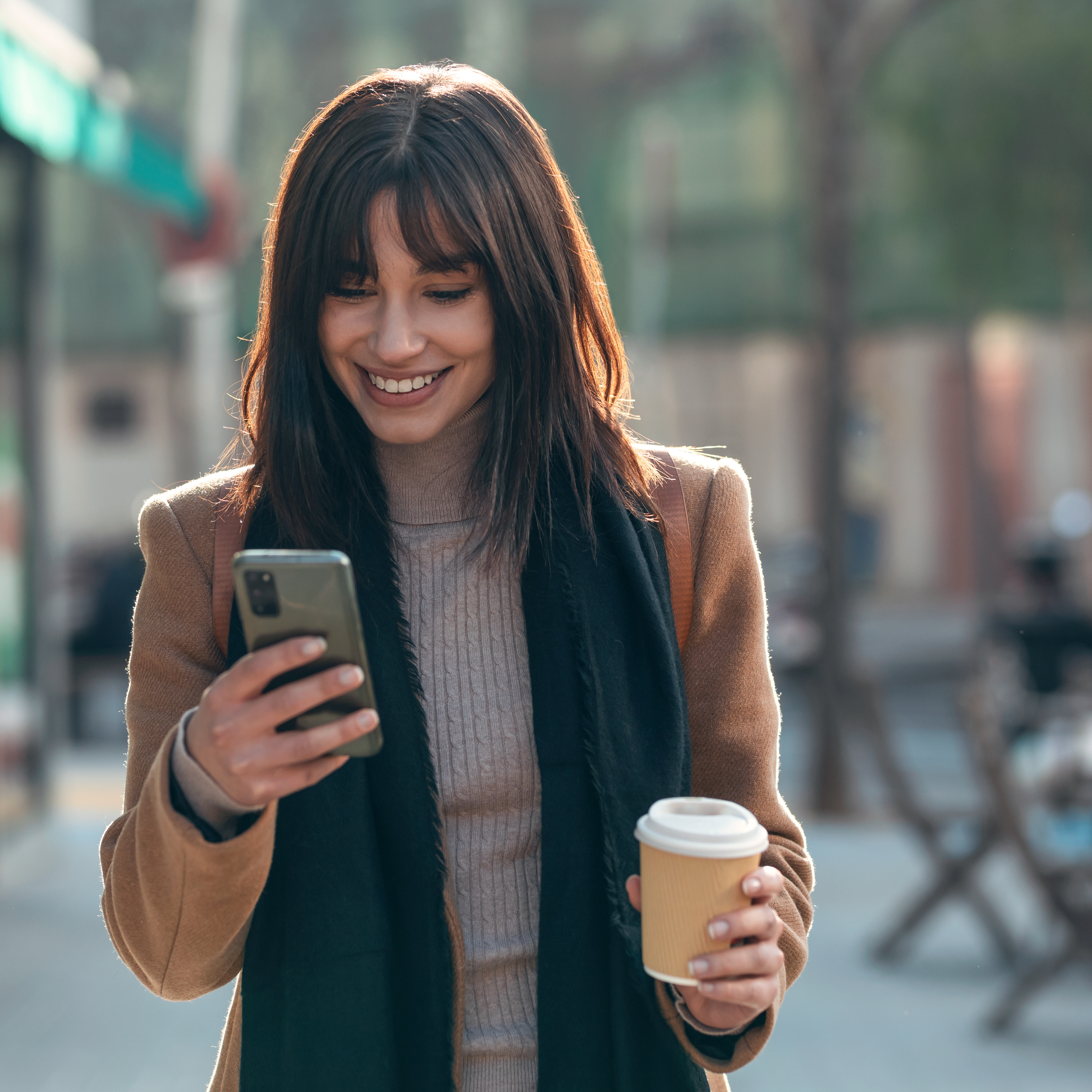 A smiling woman holds a smart phone in one hand and a coffee in the other.