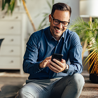 A smiling man holding a smartphone kneels on the floor.