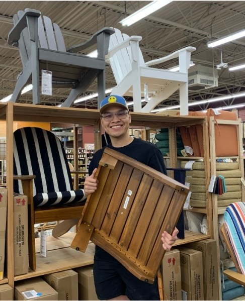 William Mai smiles while holding a wooden outdoor furniture panel inside a store, surrounded by display chairs and boxes from his family’s company, Dai Phuc Hung Thinh.