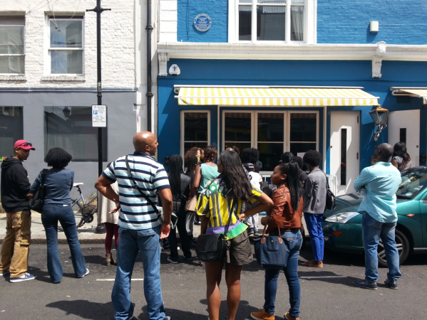 A tour group listens to a guide outside a bright blue London building with a yellow-striped awning, part of a Black History Walks event highlighting African and Caribbean heritage sites.