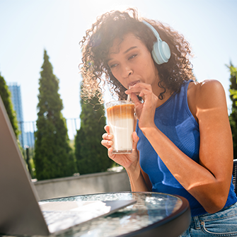 A young woman with headphones on sitting outdoors and looking at a laptop while sipping an iced latte or coffee drink through a straw.