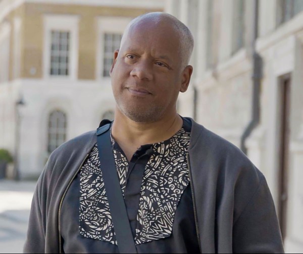 A man stands outdoors in London, wearing a patterned shirt and gray jacket, speaking during an interview for Black History Walks. The background shows historic stone buildings softly out of focus.
